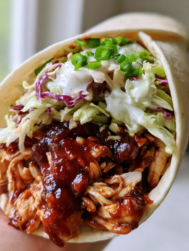 A handheld wrap at an angle featuring thick glossy dark reddish-brown BBQ sauce on chicken and opaque white creamy ranch dressing on cabbage. (BBQ Chicken Coleslaw Wraps)