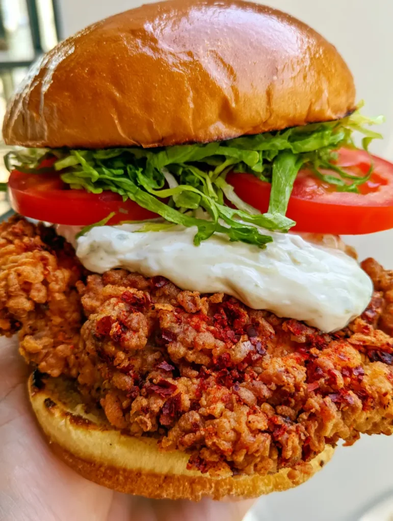 A tall Cajun Crispy Chicken Sandwich being held over a neutral background in natural daylight, showing vibrant green lettuce and red tomatoes.